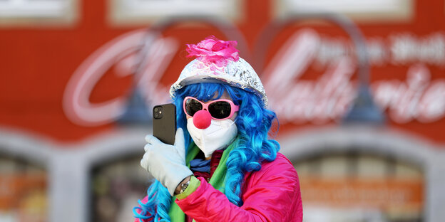Ein Clown mit Aluhut, Sonnenbrille, Atemmaske und blauer Persücke fotografiert isch selbst