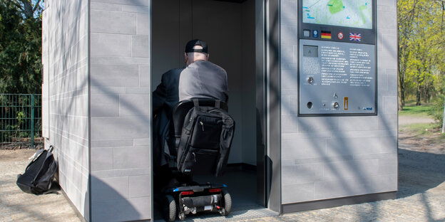 Ein graues Toilettenhäuschen ist zu sehen, in der Tür fährt ein Mensch auf einem Rollstuhl. Auf dem Toilettenhäuschen sieht man den Schatten eines Baumes, eine Stadtkarte und Informationstext, der nicht genauer zu erkennen ist.