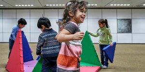 Mädchen hüpft und lacht, dahinter spielen Kinder mit bunten Schaumstoffteilen