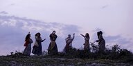 Dämmerungslicht, man sieht die Silhouetten sechs junger Frauen mit langen Röcken vor dem Himmelund Tamborin in einer Reihe