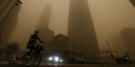 Ein Radfahrer und Autofahrer bewegen sich während der morgendlichen Hauptverkehrszeit im zentralen Geschäftsviertel von Peking inmitten eines Sandsturms an Bürogebäuden vorbei
