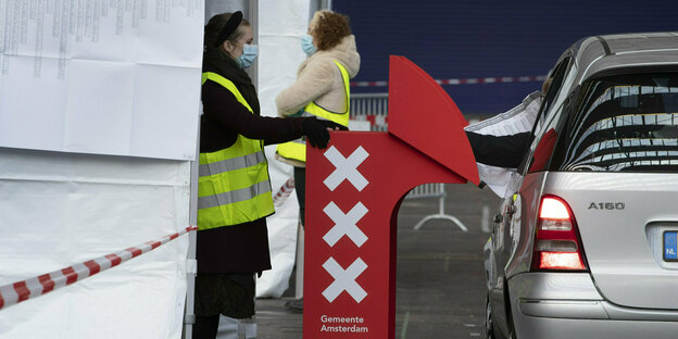 Auto parkt neben einem Zelt mit vorgebauter Wahlkabine, in der ein Mensch wählt, während ein Wahlhelfer beobachtet