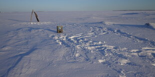 Aus einer Eisfläche die sich über das Gesichtsfeld hinaus dehnt steht ein Radio und dahinter ist ein Anzug zu sehen der an einem Holzgestell hängeden im Wind weht