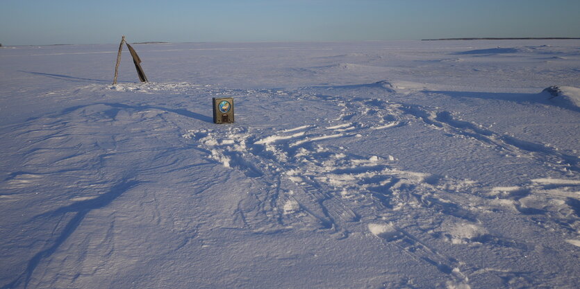 Aus einer Eisfläche die sich über das Gesichtsfeld hinaus dehnt steht ein Radio und dahinter ist ein Anzug zu sehen der an einem Holzgestell hängeden im Wind weht