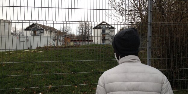 Ein Mann in weißer Jacke steht vor einem Zaun, dahinter große Gebäude