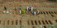 Aus der Luft aufgenommen: Zwischen Reihen ausgehobener Gräber gehen Menschen in Schutzkleidung mit einem Sarg