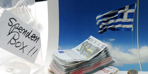 Vor einer griechischen Flagge stapeln sich Geldscheine, daneben eine Kiste mit der Aufschrift "Spendenbox".
