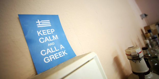 Auf einem Schrank ist ein Schild zu sehen. Aufschrift: "Bleibe ruhig, ruf einen Griechen."