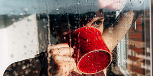 Eine junge Frau trinkt aus einer roten Tasse und schaut aus dem Fenster - Regentropfen an der Scheibe