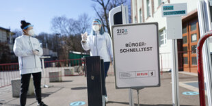 Abgesperrter Gehweg vor der Corona-Schnelltest-Station Lehrter Straße in Berlin. Zwei maskierte Mitarbeiter:innen stehen mit Abstand hinter einem Schild, auf dem "Bürger-Schnelltest" steht