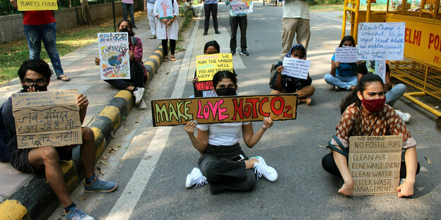 Demonstrierende sitzen mit Absatnd auf einer Straße und halten Schilder in die Höhe