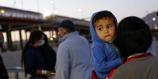 Ein Kind auf dem Arm einer Frau, im Hintergrund Grenzanlagen