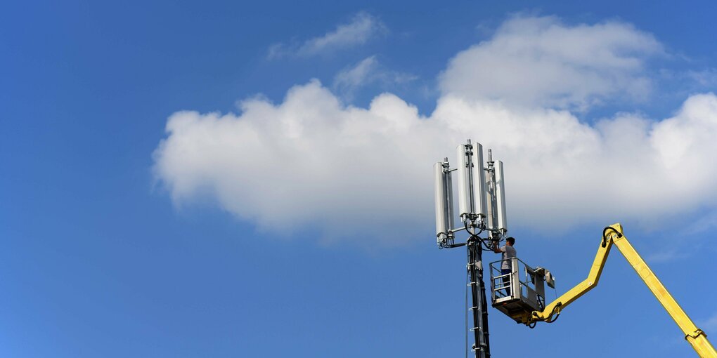 Hebebühne mit Mensch, der sich an einem UTMS-Mast vor blauem Himmel mit Wolke zu schaffen macht