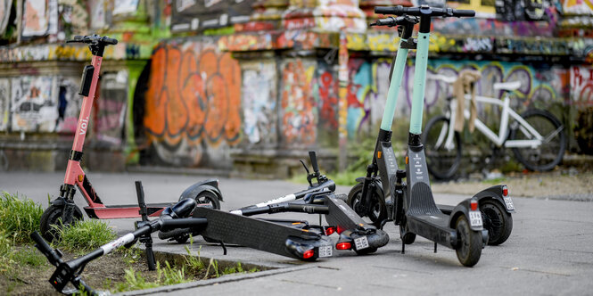 Stehende sowie kreuz und quer liegende Roller auf einem Gehsteig, daninter besprühter Fassadensockel