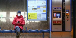 Eine Person mit Maske sitzt an einer Bushaltestelle neben einem Ticketautomaten und schaut auf ihr Handy