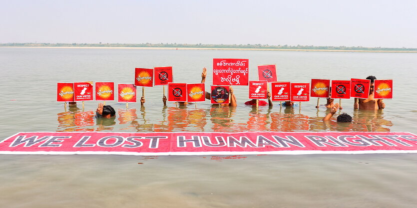 Mehrere Menschen fast untergetaucht im braunen Wasser eines Flusses. Sie halten Plakate gegen den Militärputsch in Myanmar hoch