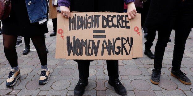 Eine Frau hält ein Plakat mit der Aufschrift "Midnight Decree = Women Angry" vor ihrem Körper. Dahinter andere Frauen, man sieht nur die untere Hälfte der Personen