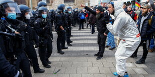 Mann mit weißem Schutzanzug wendet sich wütend gegen eine Kette behelmter Polizisten