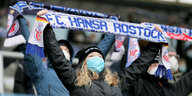 Fans mit Mundschutz halten auf der Tribüne Schals von Hansa Rostock in die Höhe