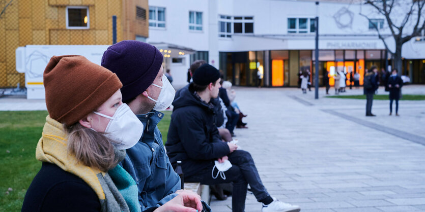 menschen mit Masken sitzen auf einer Bank vor der Philharmonie