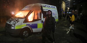 Ein Mann vor einem brennenden Polizeiwagen mit Eisenstange in der Hand skandiert