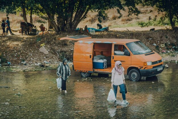 Frauen waten durchs Wasser, im Hintergrund ein orangener Kleinbus.