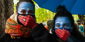Zwei Frauen mit blau bemalten Gesichtern halten sich ihre rot gefärbten Hände vor den Mund