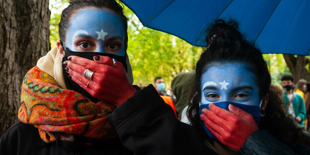 Zwei Frauen mit blau bemalten Gesichtern halten sich ihre rot gefärbten Hände vor den Mund