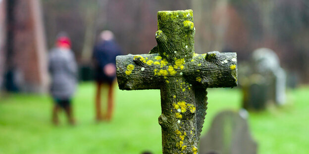 Ein Kreuz auf einem Grabstein - im Hintergrund Spaziergänger