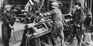4 Arbeiter*innen stehen zur Zeit des 1. Weltkrieg vor einer Maschine in einer Fabrik für Kriegsmunition