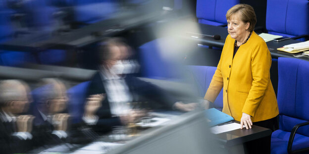 Merkel steht in gelbem Mantel vor blauem Stuhl - vor ihr in einer Spiegelung sieht man Bundestagsabgeordnete