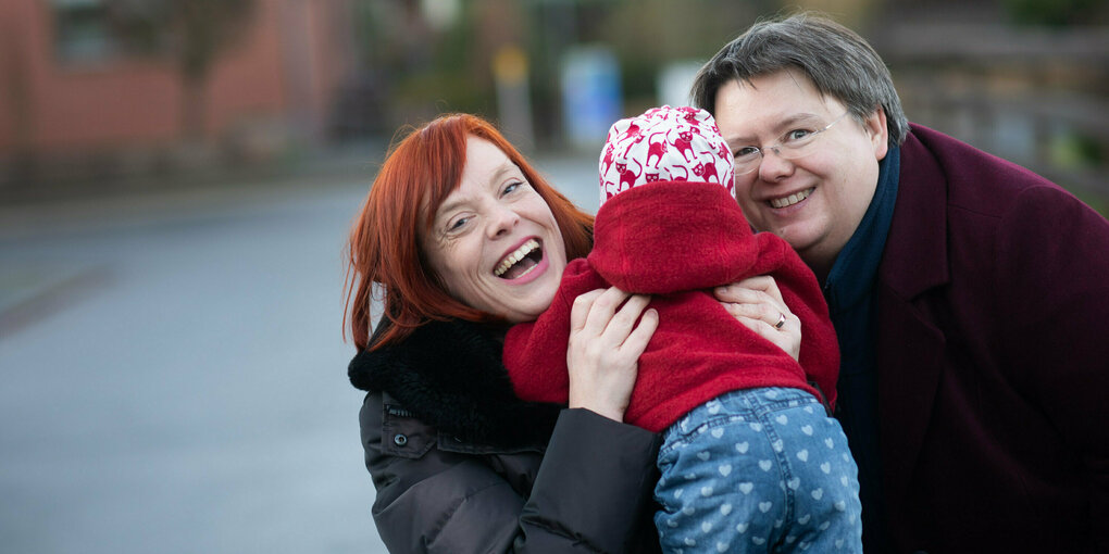 Zwei Frauen lachen, eine hebt ein Baby hoch, das von hinten zu sehen ist