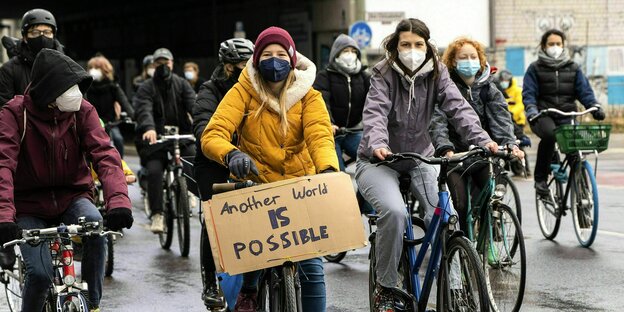 Junge Leute mit Gesichtsmasken auf einer Fahrraddemo - eine Teilnehmerin hat ein Schild: Another World is Possible