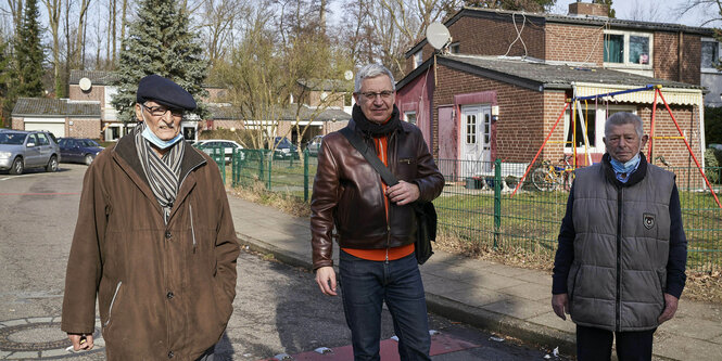 drei stehende Männer, dahinter rechts ein flaches Ziegelhaus und Bäume