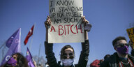 Eine Frau hält ein Plakat in die Luft auf dem auf Englisch steht: "Ich weiß, ich weiß, ich erhebe meine Stimme, ich bin so eine Bitch"
