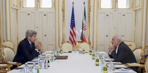 zwei Männer sitzen sich an einem gedeckten Tisch gegenüber, dahinter die Fahnen von USA und Iran