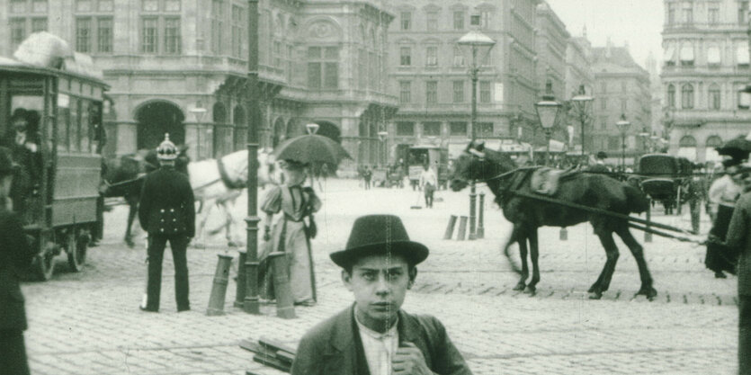 Pferdestraßenbahnen kreuzen auf dem Wiener Opernring, ein bepickelhaubter Polizist wacht auf der Verkehrsinsel, ein Junge schaut in die Kamera. Filmstill aus „Vienne, le ring“