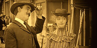 Szene aus dem schwarz-weiß Film "True Heart Suzie": Ein Mann im Anzug und eine Frau im Kleid stehen nebeneinander, der Mann hebt die Hand zum Hut