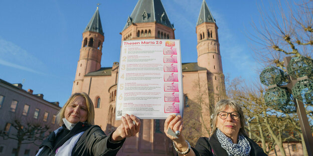 Unter blauem Himmel: Zwei Frauen halten ein Forderungsschild vor dem Mainzer Dom in die Höhe