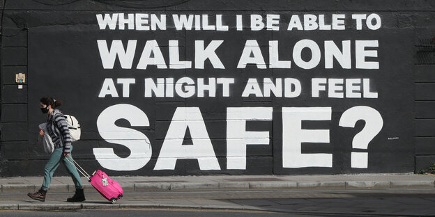 Eine Frau zieht ihren Rollkofer an einem Wandbild vorbei, dessen Text lautet: "When will I be able to walk alone at night and feel safe?"