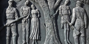 Ein Relief zeigt einen Baum, an dem drei Afrikaner:innen erhängt worden sind, davor zwei Soldaten mit Gewehren
