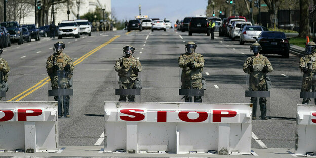 Mehrere bewaffnete Sicherheitskräfte stehen hinter einer Straßenbarriere auf einer breiten Allee