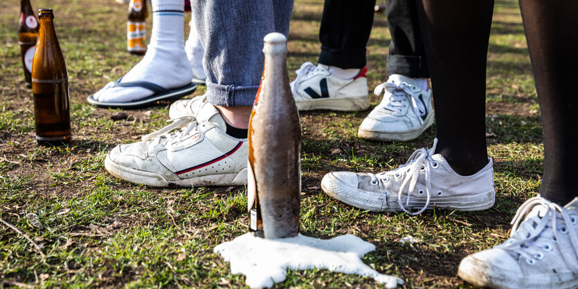 Es sind einige Fußpaare zusehen, die alle weiße Sneaker tragen. Vor ihnen stehen geöffnete Bierflaschen, manche schäumen über