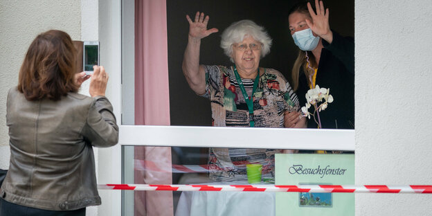 Eine alte Mann und eine Pflegerin stehen hinter einem Glasfenster und winken einer Person auf der anderen Seite zu