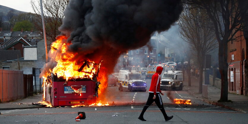 Ein Mann läuft über eine Straße auf der ein roter Bus in Flammen steht