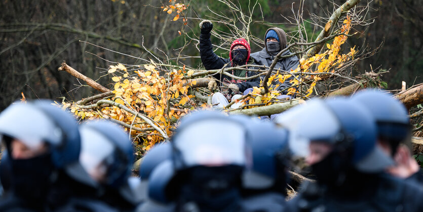 Zwei bunt vermummte Personen umringt von Laub und Zweigen, davor behelmte Polizei