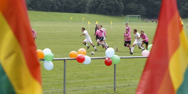 zwei Teams spielen Fußball, im Vordergrund zwei Regenbogenfahnen