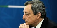 Mario Draghi hält bei einer Pressekonferenz seine Maske in der Hand und blickt konzentriert nach links