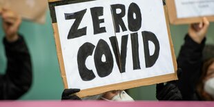 Eine Person hält ein Schild in die Höhe auf dem mit schwarzer Schrift geschrieben steht "Zero Covid"