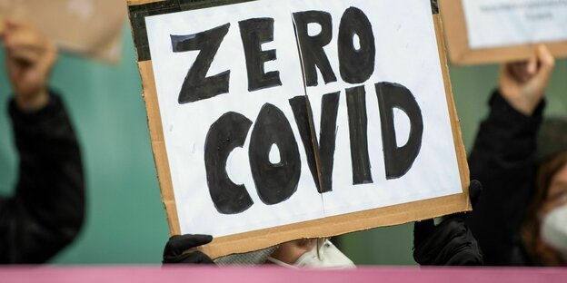 Eine Person hält ein Schild in die Höhe auf dem mit schwarzer Schrift geschrieben steht "Zero Covid"
